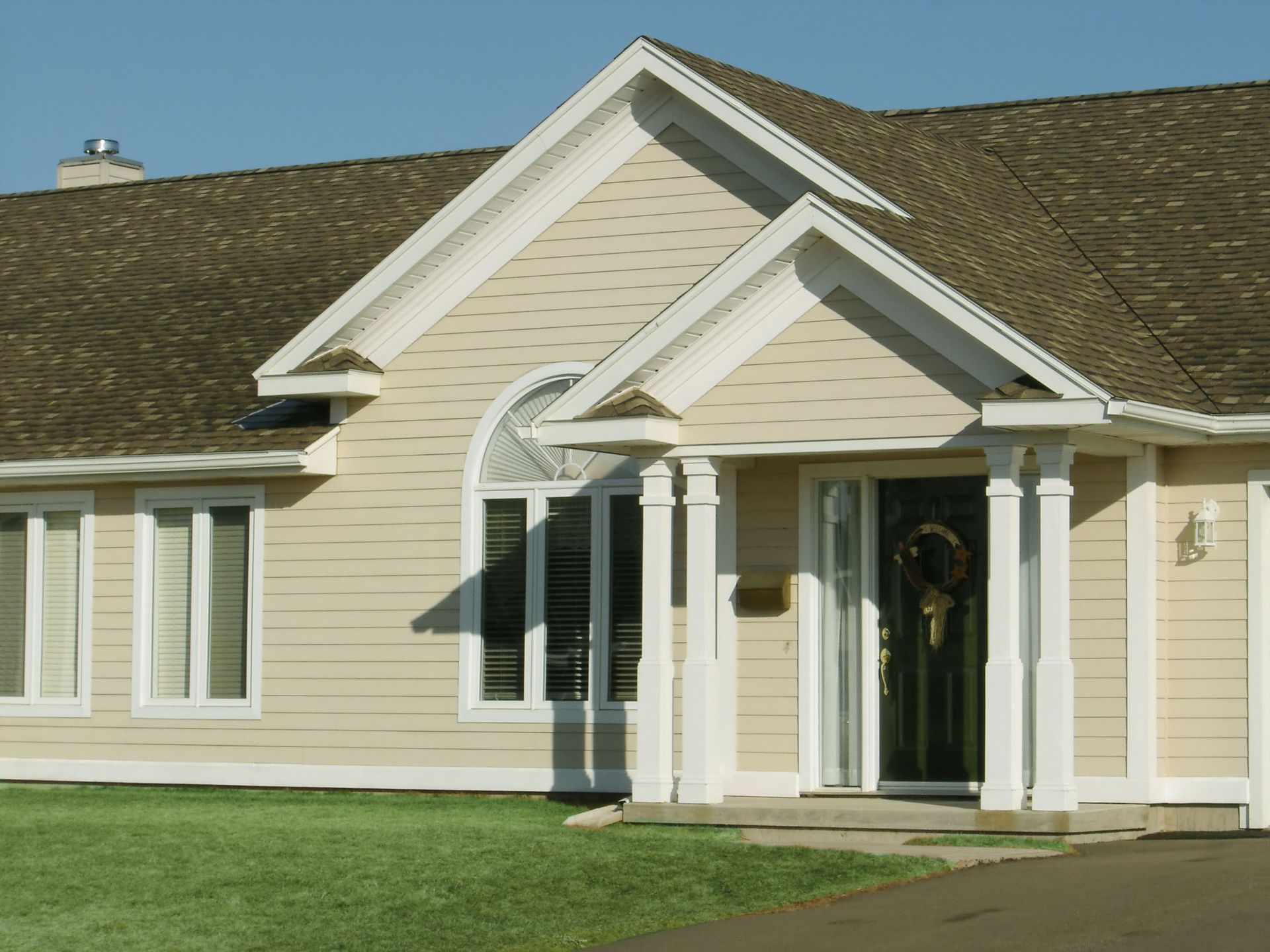 A single-story, light yellow house with a gabled porch entrance, white columns, and a brown shingled roof under a blue sky.